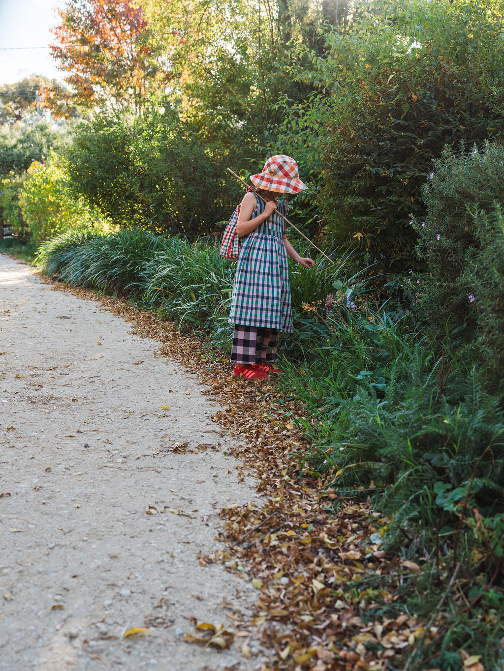 A child pausing on a bush path in checked Cousin Pants, flowers in hand and a stick over her shoulder