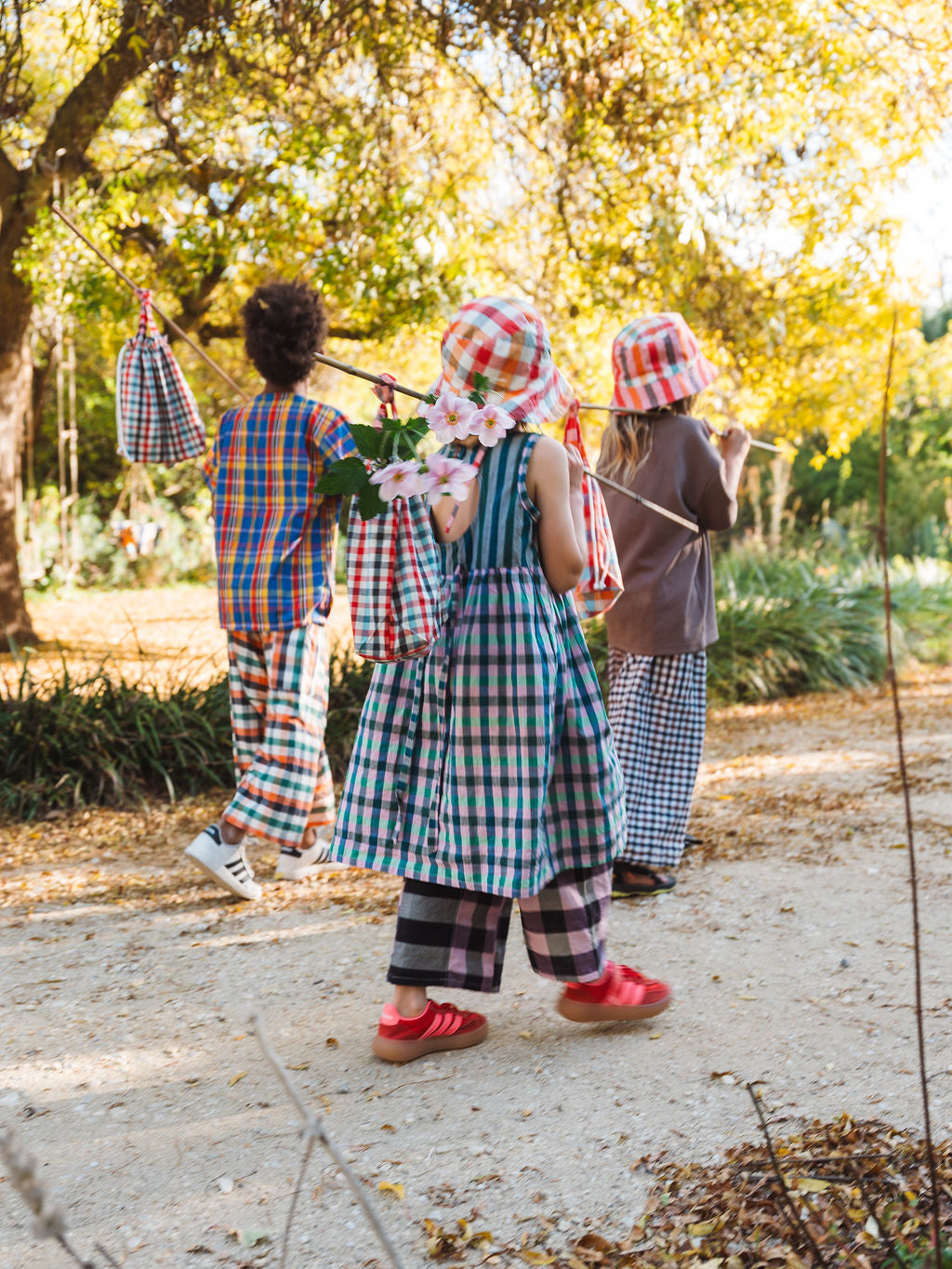 Three children walking a bush track in checked Cousin Pants, bundles slung on sticks